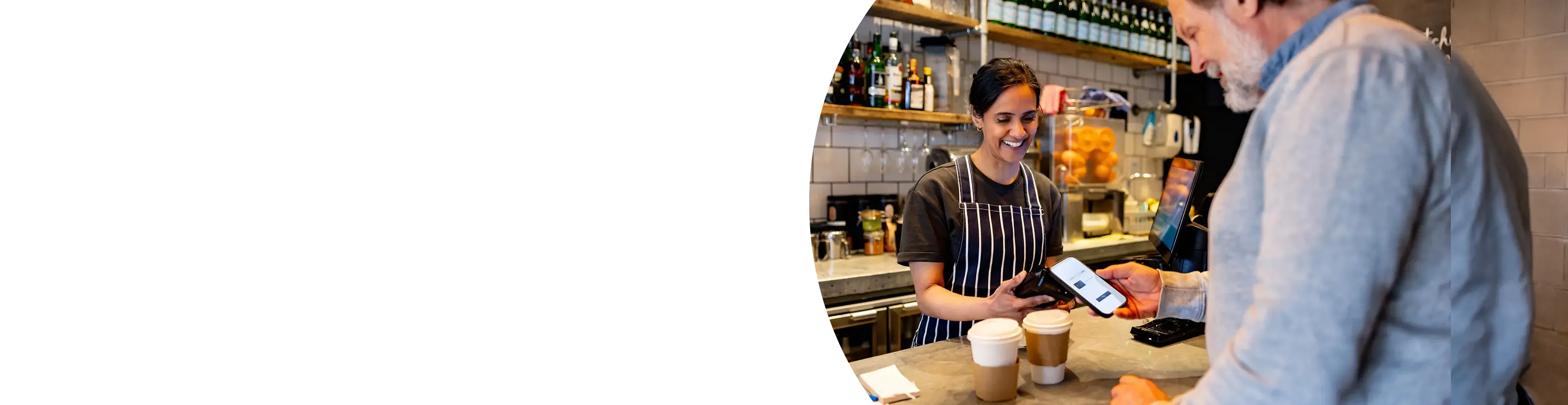 A smiling barista holds a card reader as a customer pays with his phone at a cafe counter.